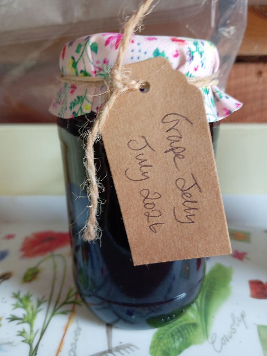 A jar of grape jelly with a floral fabric top and a handwritten brown label reading Grape Jelly July 2026