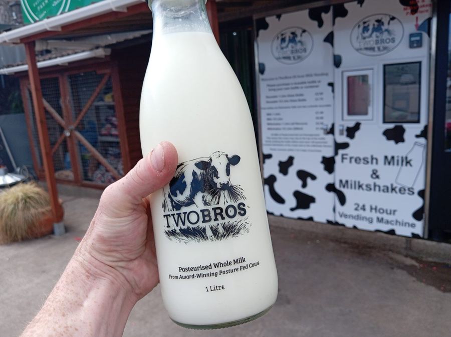 A glass litre bottle of Two Bros pasteurised whole milk held up in front of the 24-hour milk vending machine at Easter Compton Farm Shop